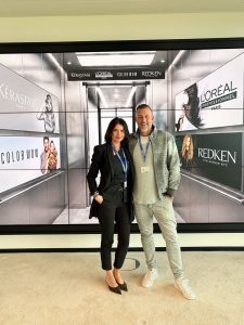 Marjam_Laurentius_Robert_Color_Wow_Launch - Friseur Bonn ❤️ Laurentius Excellence in Hair ⭐ Two professionally dressed people pose together (woman in black suit, man in gray jacket) in front of a wall of haircare brand ads (L'Oréal, Redken, Color Wow).