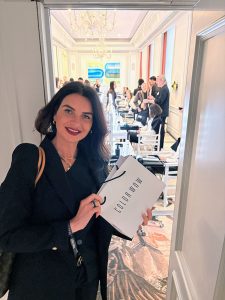 Marjam_Laurntius_Color_Wow - Friseur Bonn ❤️ Laurentius Excellence in Hair ⭐ Smiling woman in a black outfit stands at a doorway, holding a Color Wow shopping bag in a chandelier-lit event room with people in the background.